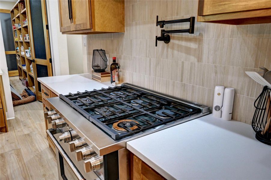 This kitchen features a modern gas stove with a sleek design, ample counter space, and warm wood cabinetry. It includes a convenient pot filler and leads into a spacious pantry with sliding doors, offering plenty of storage.