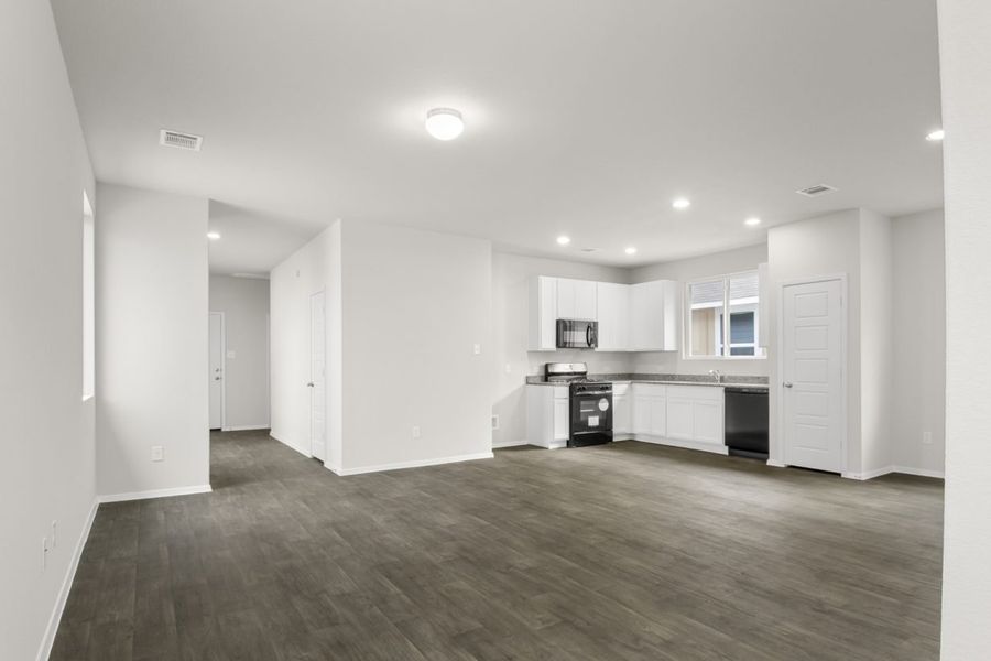 Image of living room with brown flooring and light grey walls with a kitchen in the distance
