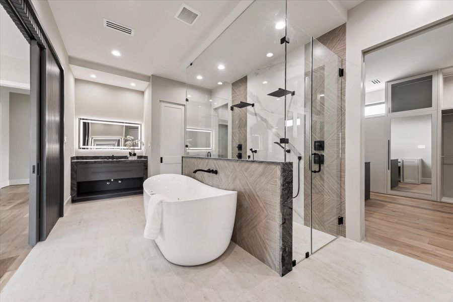 This spa-like primary bathroom is designed to impress with its modern elegance and luxurious finishes. At the center, a freestanding soaking tub creates a striking focal point, perfectly framed by a glass-enclosed shower featuring dual rainfall showerheads and dramatic tile detail.