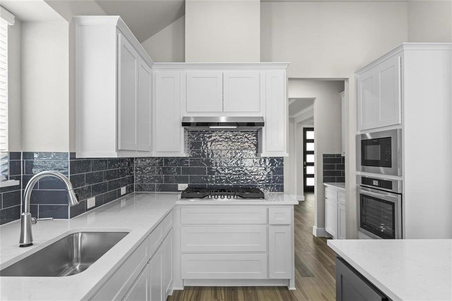 Kitchen featuring white cabinetry, light-toned countertops, dark tile backsplash, stainless steel appliances, and wood-finish flooring Kitchen featuring white cabinetry, light-toned countertops, dark tile backsplash, stainless steel appliances, and wood-finish flooring