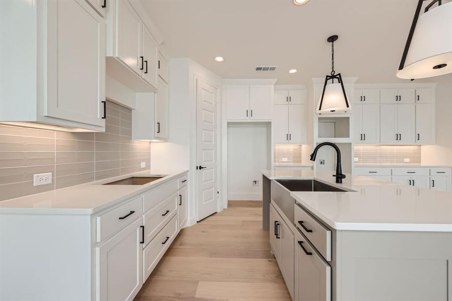 Kitchen with light wood-type flooring, tasteful backsplash, an island with sink, white cabinets, and decorative light fixtures