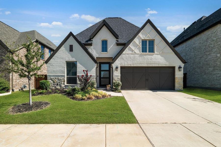 French country style house featuring brick siding, a front lawn, concrete driveway, a shingled roof, and stone siding French country style house featuring brick siding, a front lawn, concrete driveway, a shingled roof, and stone siding