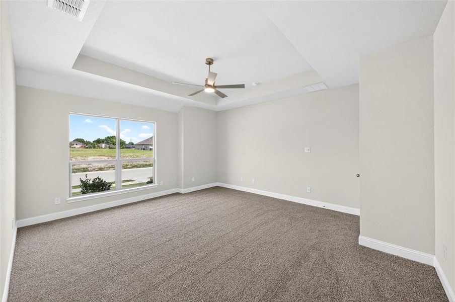 Spacious interior room featuring a tray ceiling with a ceiling fan, light gray wall paint, gray carpet flooring, and a large window providing ample natural illumination