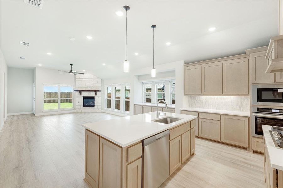 Open kitchen with center island, light wood cabinets, and pendant lights, open to living room with fireplace.