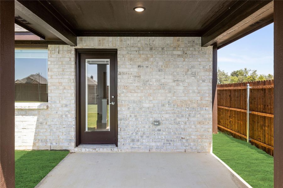 Covered back patio with concrete floor, dark entry door, light brick exterior, and wood privacy fence surrounding green lawn.