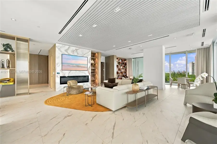 Open-concept interior featuring large-format tile flooring, a floor-to-ceiling stone-clad fireplace wall, custom built-in shelving, a tiered slatted ceiling with recessed lighting, and floor-to-ceiling windows Open-concept interior featuring large-format tile flooring, a floor-to-ceiling stone-clad fireplace wall, custom built-in shelving, a tiered slatted ceiling with recessed lighting, and floor-to-ceiling windows