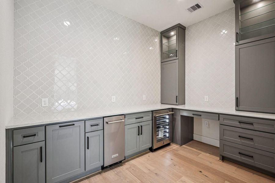This photo showcases a modern wine room with sleek gray cabinetry, a stylish patterned tile backsplash, and light wood flooring. It features ample counter space, a built-in wine cooler, and contemporary lighting, creating a functional and elegant space.