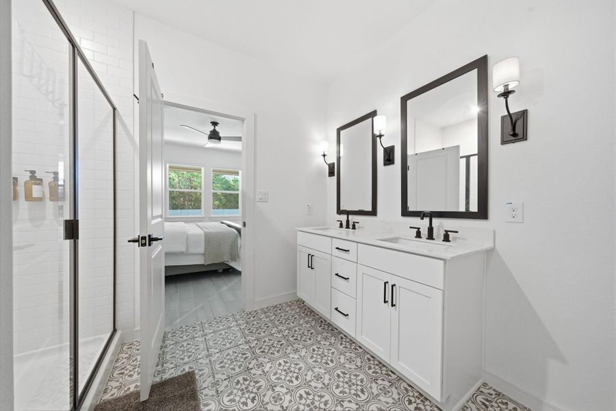 Bright, modern bathroom with elegant patterned tile flooring, dual sinks, and large mirrors. It connects to a bedroom with a view of greenery through multiple windows.