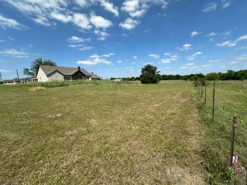 Expansive open lot featuring a partial wire fence and a distant stone-clad dwelling with a dark shingle roof and a covered porch