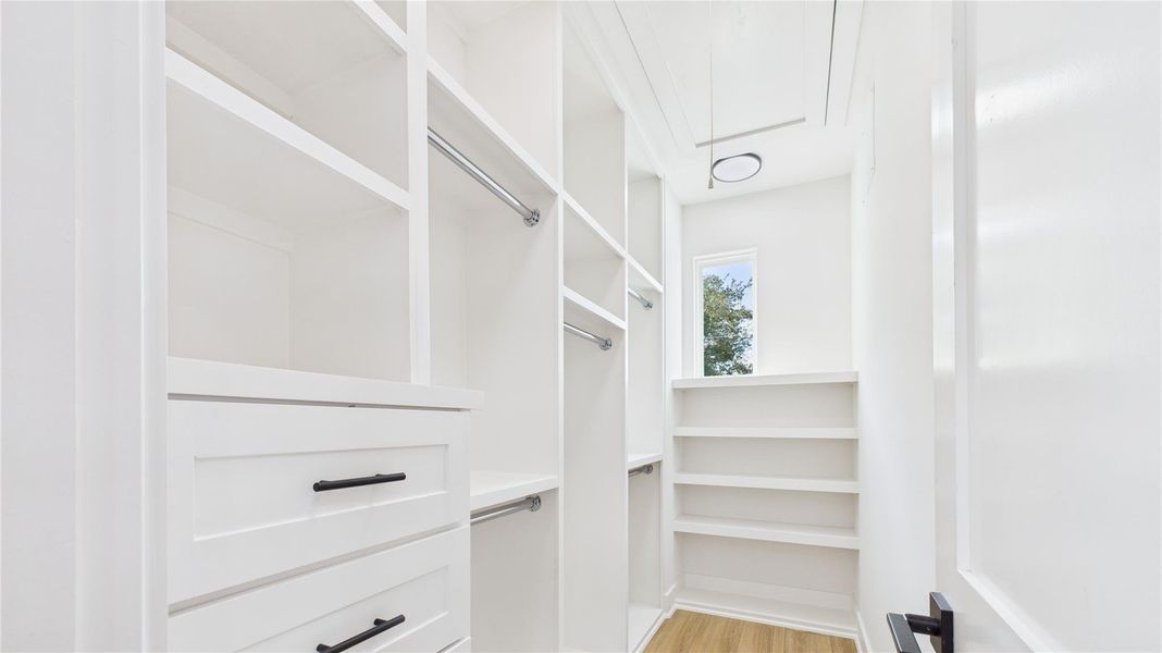 This photo showcases a bright, modern walk-in closet with ample storage. It features white shelving, hanging rods, drawers, and a small window for natural light, complemented by a light wood floor.