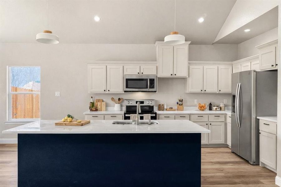 Two tone kitchen with a center island with sink, hanging light fixtures, light wood-type flooring, light stone countertops, and vaulted ceiling
