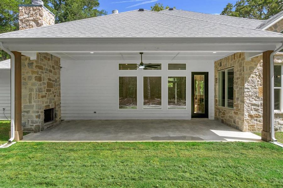 Back of house featuring stone siding, a patio, a shingled roof, a stone fireplace, and a yard Back of house featuring stone siding, a patio, a shingled roof, a stone fireplace, and a yard