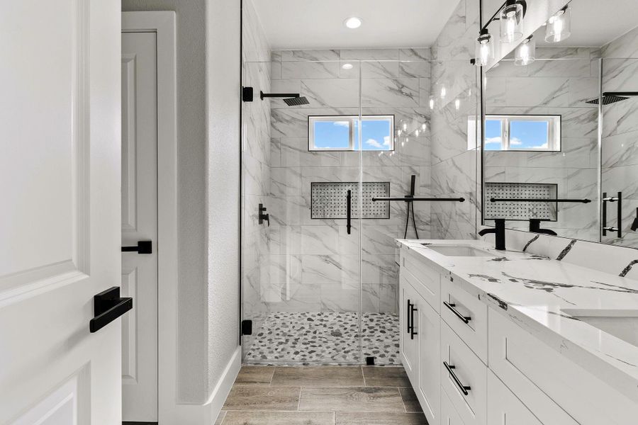 This modern bathroom features a spacious glass-enclosed shower with dual rain showerheads and pebble flooring. The vanity boasts a sleek marble countertop with ample storage. Bright natural light filters through small windows, enhancing the elegant marble tile walls.
