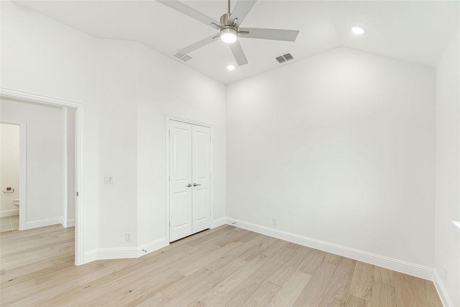 Empty bedroom with light wood floors, ceiling fan, double closet doors, and adjoining bathroom