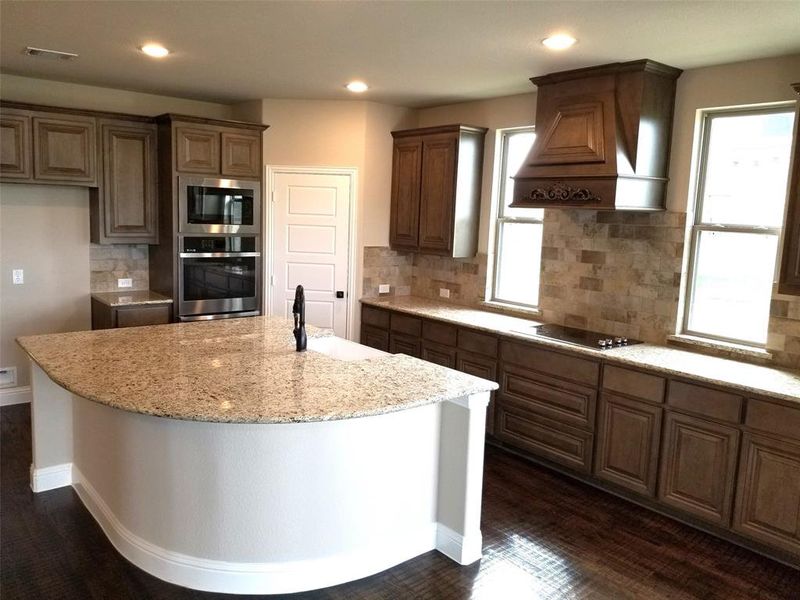 Kitchen with windows, decorative tile backsplash, stainless steel built-in double oven & cooktop, and custom cabinet vent hood to outside