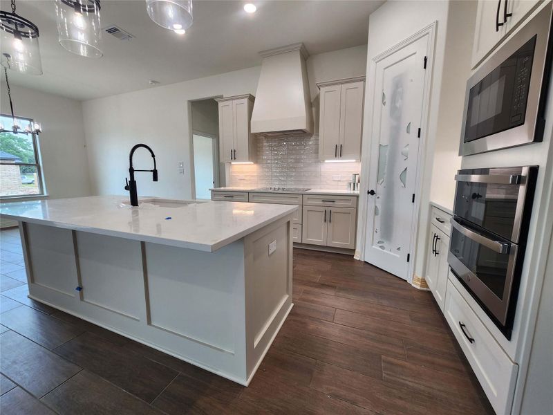 Kitchen featuring an expansive island with a white countertop and integrated sink, stainless steel built-in double ovens, light-toned cabinetry, a subway tile backsplash, and wood-finish flooring