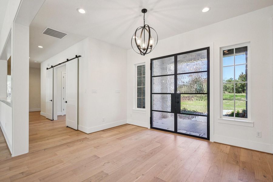 This photo showcases a bright entryway with large glass doors and side windows, offering ample natural light. It features a modern chandelier, light wood flooring, and clean white walls, providing a fresh and inviting atmosphere. A sliding barn door adds a stylish touch.