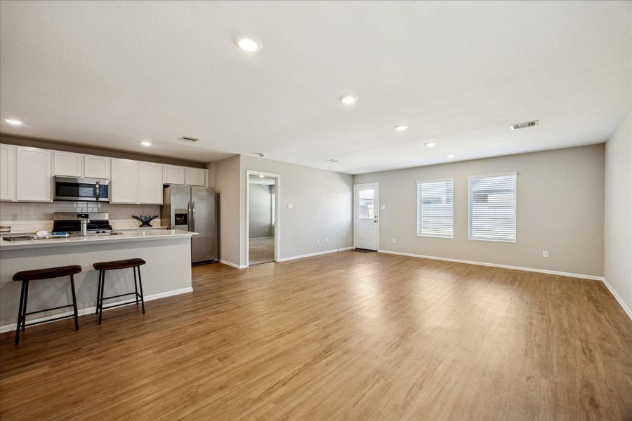 This spacious open-concept living area features a modern kitchen with stainless steel appliances and a large island with seating. The room is bright with natural light from multiple windows, and the vinyl  flooring adds warmth and elegance.