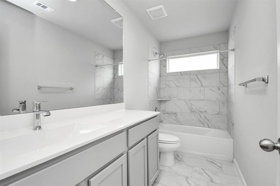 Secondary bathroom, where the tile flooring harmonizes with a bath/shower combo surrounded by tasteful tile. Light wood cabinets, coupled with a gorgeous countertop, elevate the space, cultivating a truly welcoming atmosphere. Sample photo of completed home with same plan. Actual colors and selections may vary.