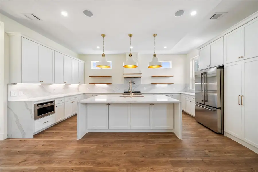 Spacious kitchen features a modern design with white cabinetry, marble countertops, and a large island. It includes stainless steel appliances, pendant lighting, and ample natural light, complemented by wood floors.