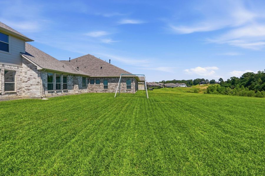 This photo showcases a spacious backyard with a well-maintained lawn, perfect for outdoor activities. The home features a brick exterior with multiple windows, providing a view of a serene landscape and neighboring houses in the distance.