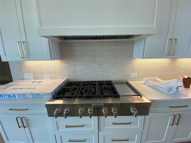 Kitchen view of premium range hood, stainless steel gas stovetop, backsplash, white cabinets, and light stone counters