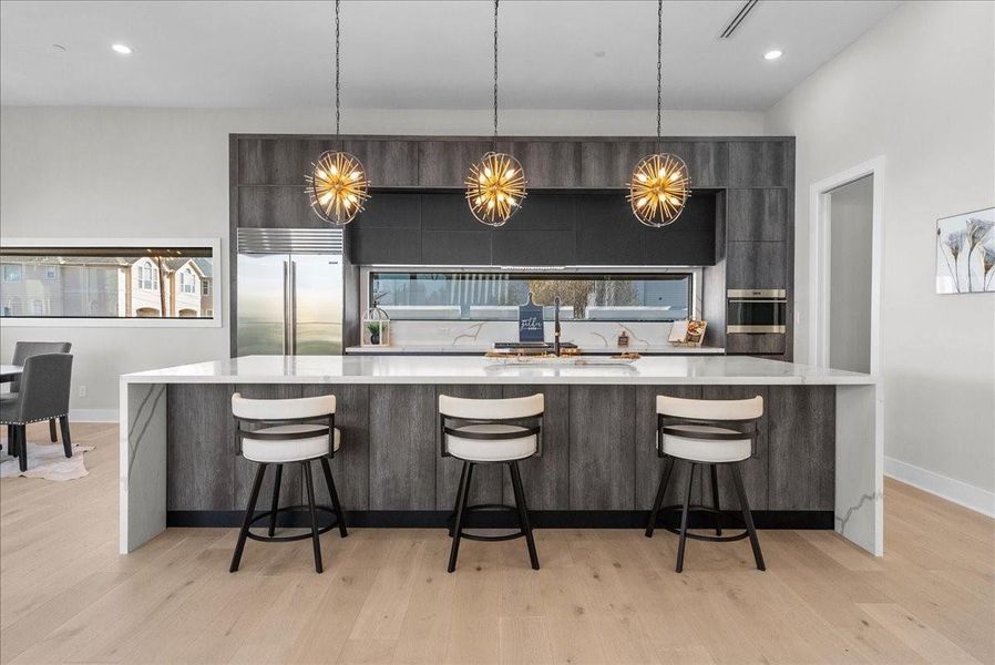 This modern kitchen features a spacious island with seating for three, sleek dark cabinetry, and high-end stainless steel appliances. Elegant pendant lights add a stylish touch, while the open layout connects to the dining area, enhancing the airy feel. This modern kitchen features a spacious island with seating for three, sleek dark cabinetry, and high-end stainless steel appliances. Elegant pendant lights add a stylish touch, while the open layout connects to the dining area, enhancing the airy feel.