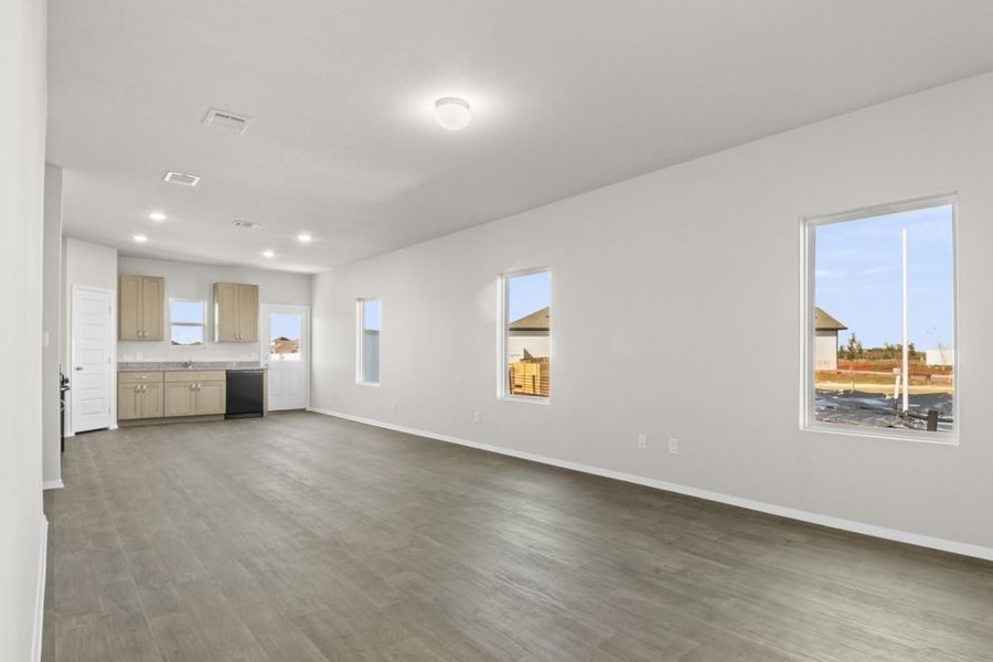 Image of a living room with dark vinyl flooring, light grey walls, windows and a kitchen in the distance