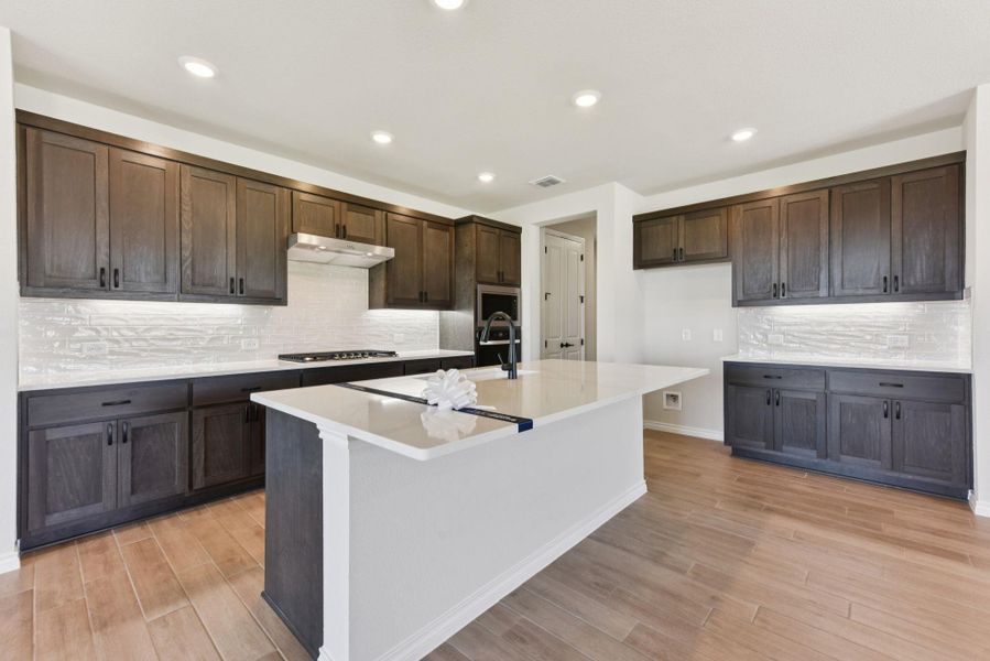 Kitchen featuring backsplash, dark brown cabinetry, recessed lighting, light stone countertops, and an island with sink