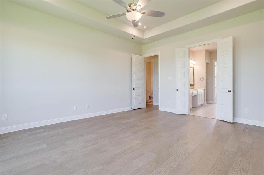 Unfurnished bedroom featuring light wood-type flooring, a raised ceiling, connected bathroom, and ceiling fan