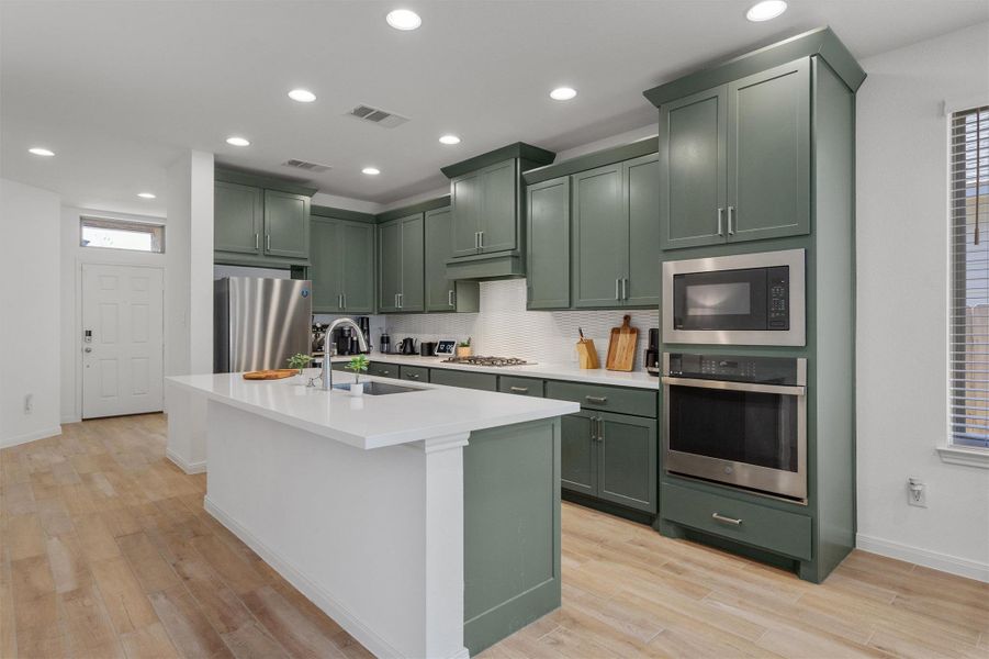 Kitchen with green cabinetry, stainless steel appliances, an island with sink, light wood finished floors, and recessed lighting