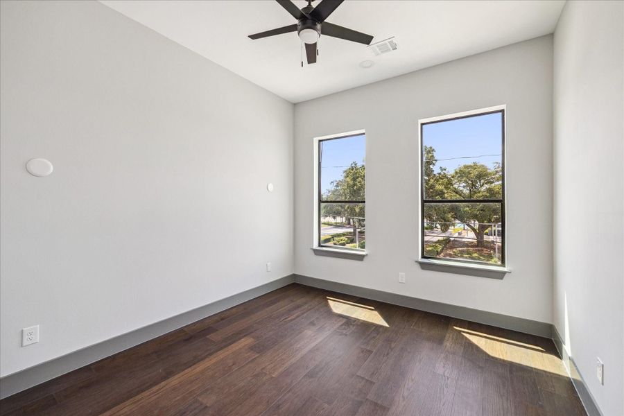 Wood floors, a ceiling fan, and wide windows shape the secondary bedroom, where abundant light and a spacious layout make it equally suited for restful nights or a home office setup.