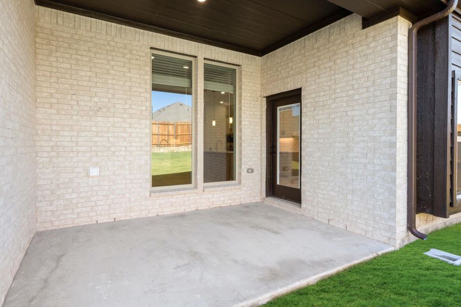 Covered back patio with concrete floor, white brick exterior, large window, and dark-framed door leading inside