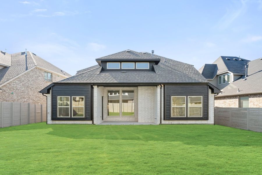 Rear exterior of single-story home with dark siding, white brick, large windows, and fenced backyard with green lawn.