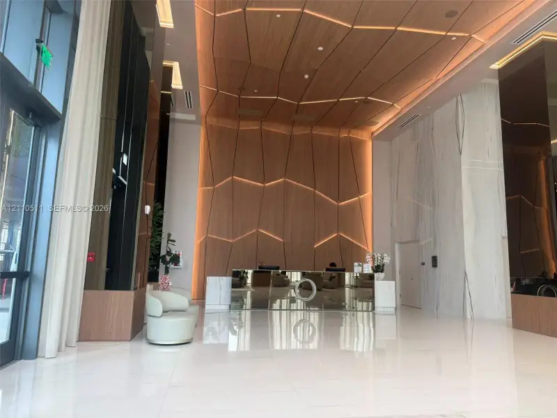 Lobby featuring high ceilings, large-format tile flooring, a geometric wood-paneled accent wall with integrated lighting, full-height stone cladding, and floor-to-ceiling glass paneling