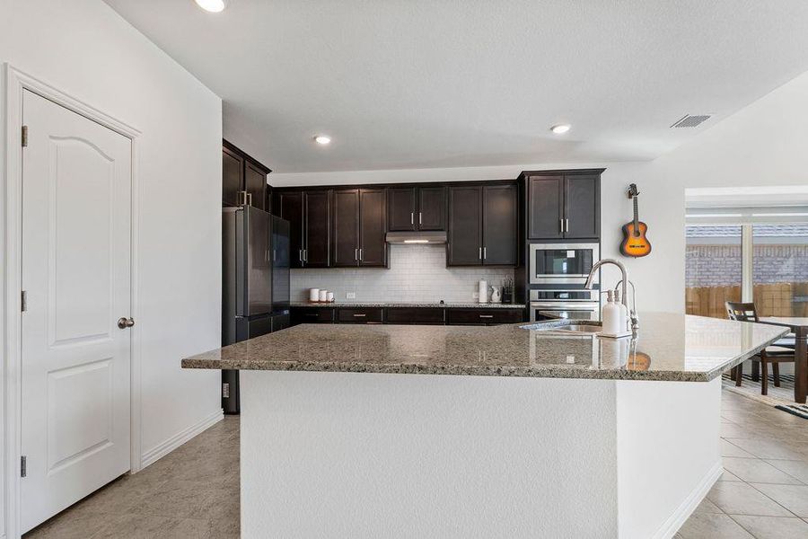Kitchen featuring dark wood finish cabinets, stainless steel appliances, a center island with sink, tasteful backsplash, and dark stone counters Kitchen featuring dark wood finish cabinets, stainless steel appliances, a center island with sink, tasteful backsplash, and dark stone counters