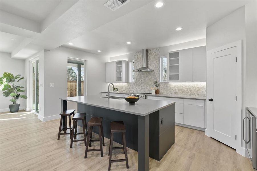 Kitchen featuring white cabinetry, light hardwood / wood-style floors, a kitchen island with sink, wall chimney range hood, and sink