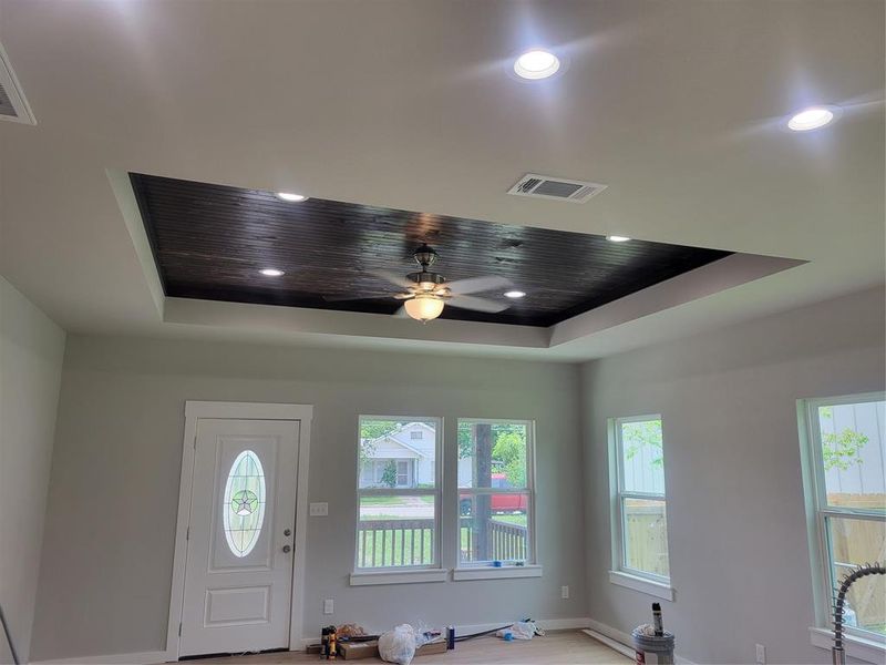 Tray ceiling with dark wood-finish planking and recessed lighting, contemporary ceiling fan, white paneled entry door with oval leaded glass insert, multiple double-hung windows, and light wood-finish flooring