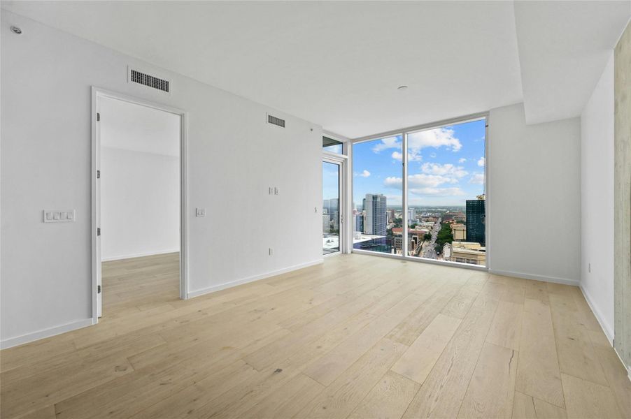 Step into this sleek and contemporary bedroom featuring floor-to-ceiling windows that frame spectacular views of the city and beyond. The natural light floods the room, enhancing the airy ambiance created by the light wood flooring and neutral color palette. The exposed concrete accent wall adds an industrial touch to the minimalist design.