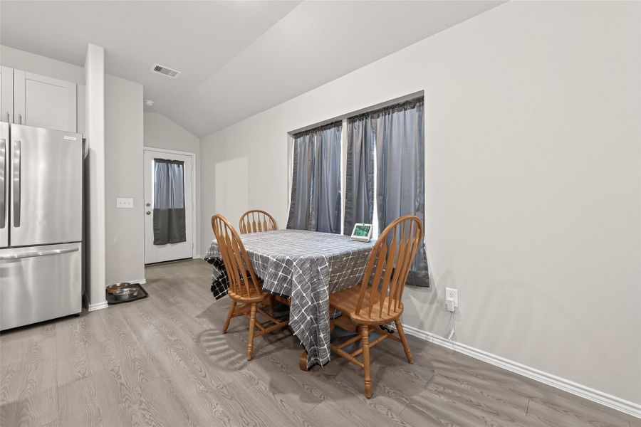 Bright dining area with a modern feel, featuring a table set for four. The space has light wood flooring, a stainless steel refrigerator, and a door leading outside. Perfect for cozy meals and gatherings.