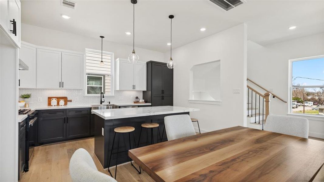 This modern kitchen features sleek black and white cabinetry, a central island with barstools, and pendant lighting. The open layout flows into a dining area with a wooden table, and a staircase leads to the upper level. Large windows provide natural light, creating a bright and inviting space.