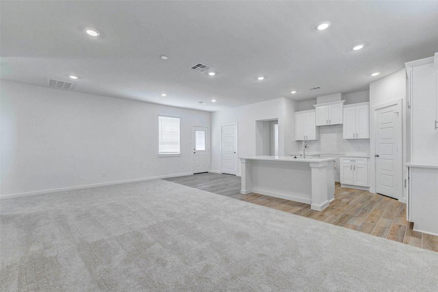 Open-concept living, dining, and kitchen area with soft carpet in the living space, wood-look tile in the kitchen, recessed lighting, white cabinetry, and a large island. The spacious layout offers flexibility for everyday living, entertaining, and comfortable furniture placement.