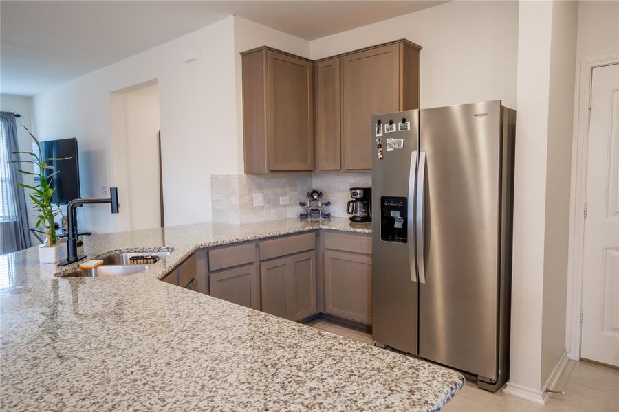 Kitchen featuring granite-style countertops, under-mount sink with a matte black faucet, light grey cabinetry, tile backsplash, and stainless steel refrigerator Kitchen featuring granite-style countertops, under-mount sink with a matte black faucet, light grey cabinetry, tile backsplash, and stainless steel refrigerator