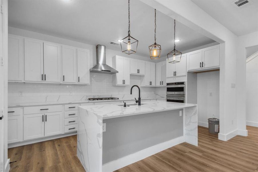 Kitchen with wall chimney range hood, an island with sink, white cabinetry, decorative light fixtures, and sink