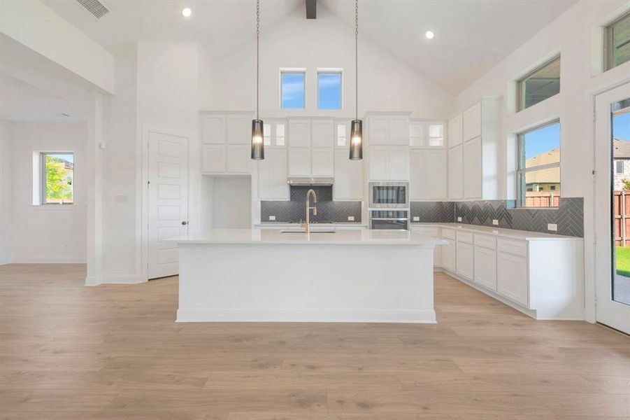 Modern kitchen featuring a large central island, light wood-finish flooring, white cabinetry, dark herringbone-patterned backsplash, and stainless steel built-in appliances Modern kitchen featuring a large central island, light wood-finish flooring, white cabinetry, dark herringbone-patterned backsplash, and stainless steel built-in appliances