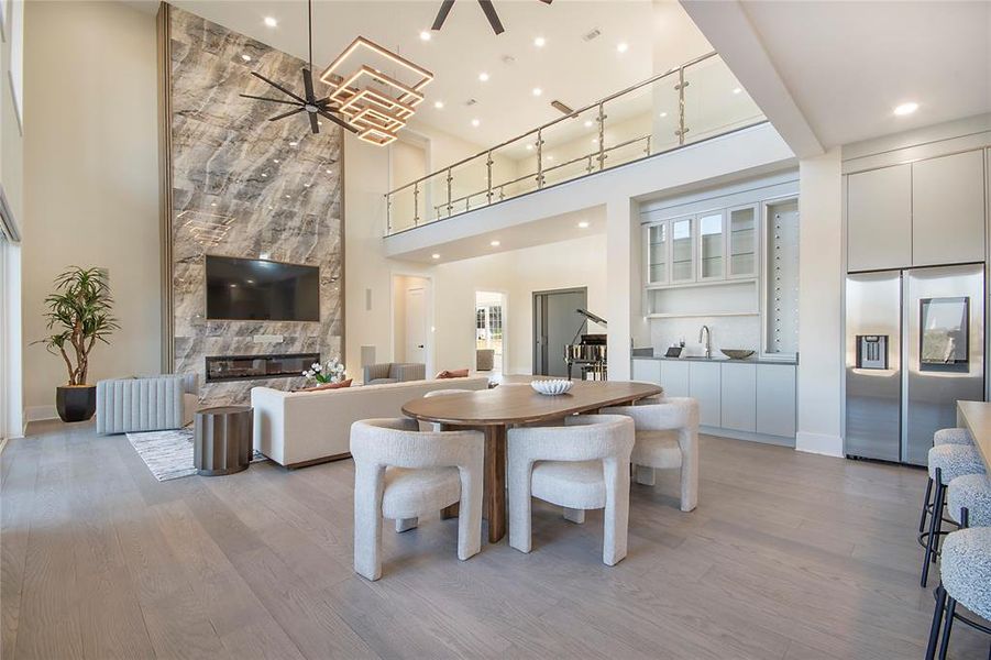 Dining space featuring ceiling fan, light wood-style flooring, a fireplace, and a high ceiling Dining space featuring ceiling fan, light wood-style flooring, a fireplace, and a high ceiling