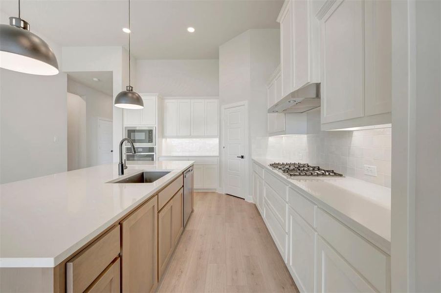 Modern kitchen featuring a spacious island with an undermount sink and matte black faucet, light wood-finish flooring, white cabinetry, built-in stainless steel appliances, and recessed lighting