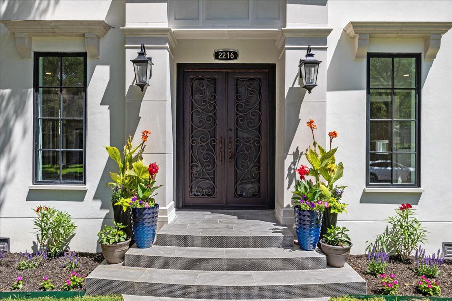 This elegant front entrance with dark iron ornate doors are framed by large widows and stylish lanterns.  The area is beautifully landscaped with colorful potted plants, creating a welcoming and sophisticated curb appeal.