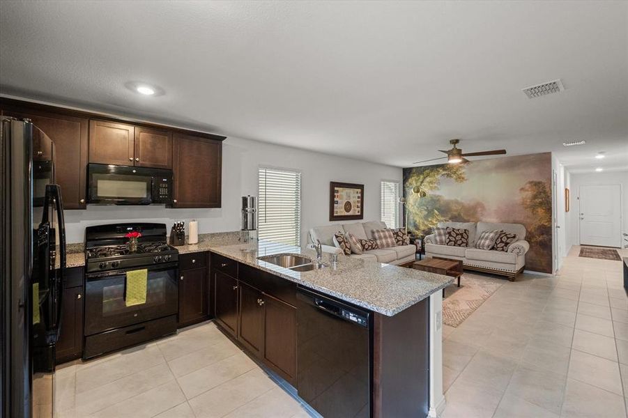 Kitchen featuring dark brown cabinetry, black appliances, light stone countertops, open floor plan, and light tile patterned flooring
