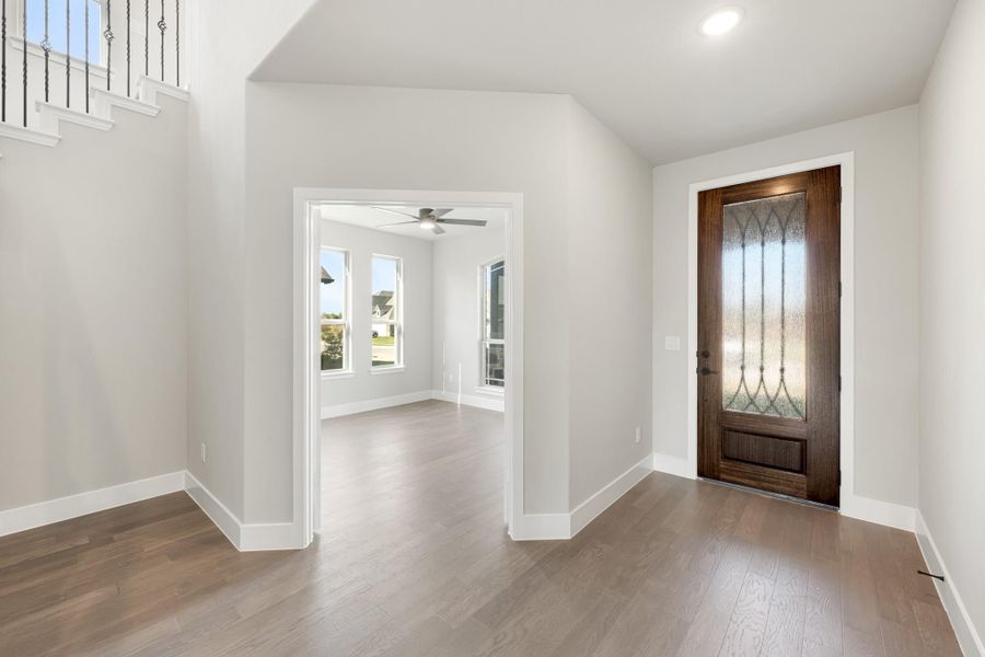 Entryway with dark wood front door featuring decorative glass panel and hardwood floors, opening to adjacent room with ceiling fan.